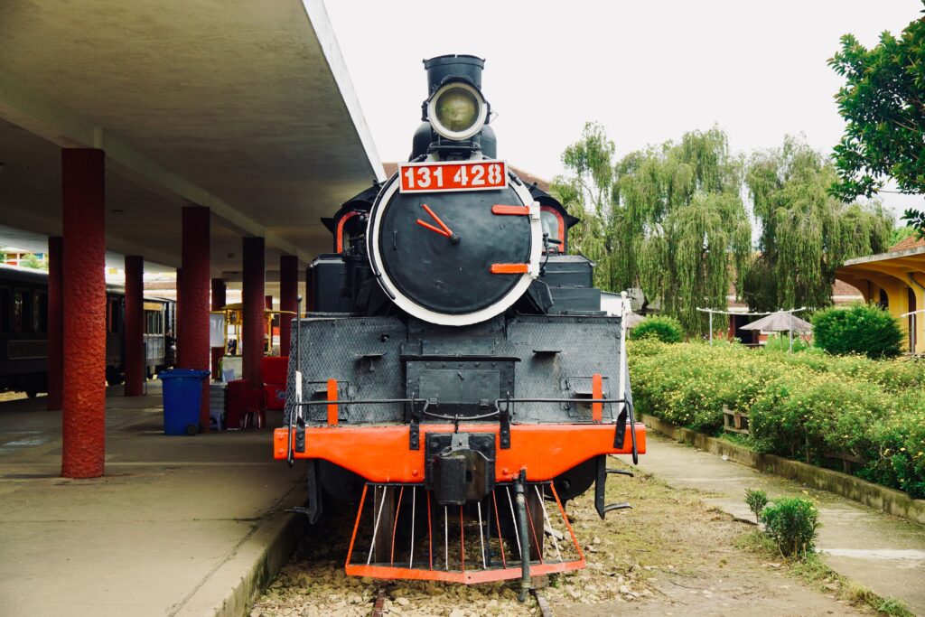Front view of a vintage steam train at an outdoor station with greenery.
